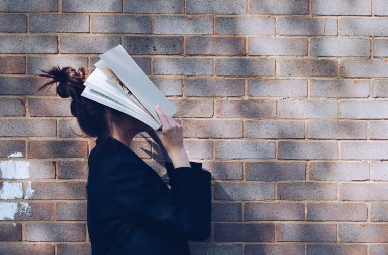 A girl is standing in front of a brick wall and holding a book over her face.