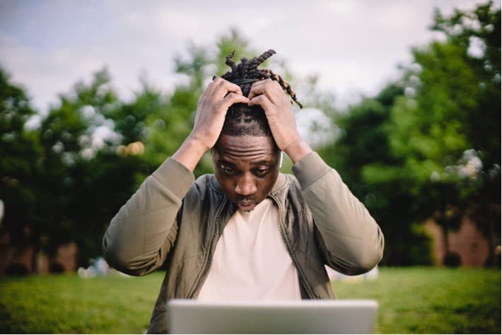 A man sitting at a laptop outdoors and holding his head with both hands in confusion