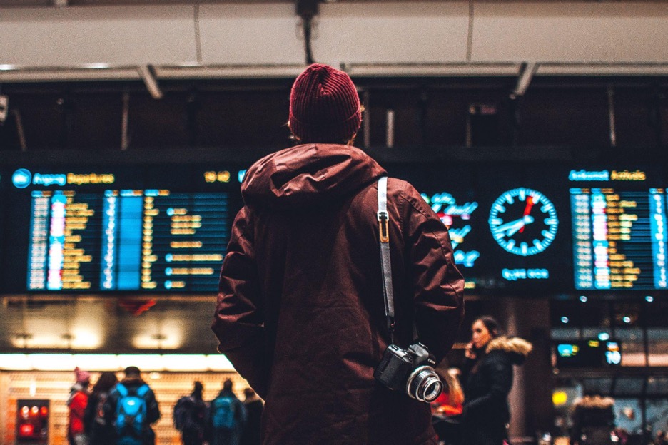 A man wearing a jacket and a hat is looking at the departures at an airport