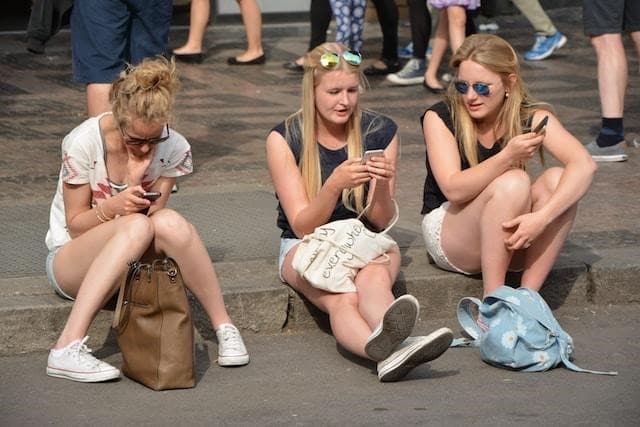 Three girls sitting on the sidewalk, using their smartphones.