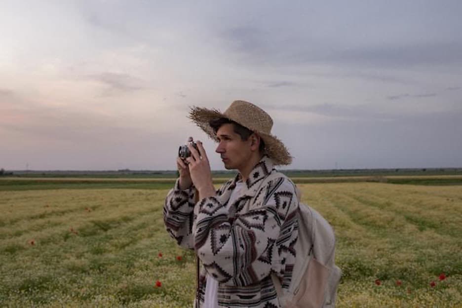 A young man holding a camera with a straw hat in a meadow