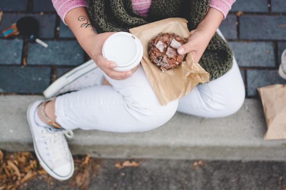 A woman sitting on the sidewalk holding a browny and a takeaway coffee