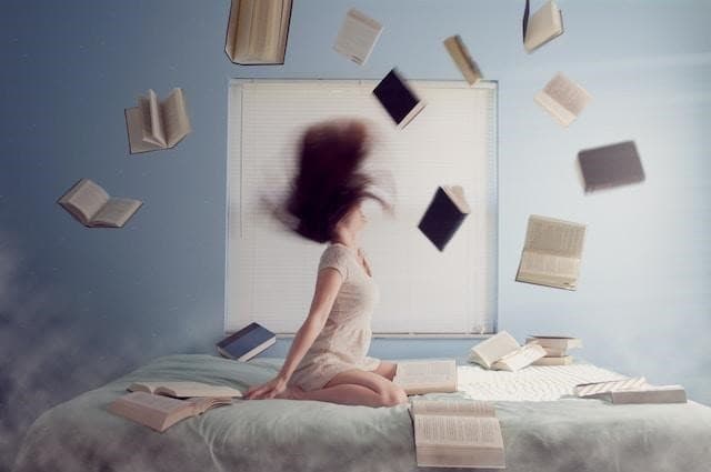 A woman sitting on the bed with books flying around her.