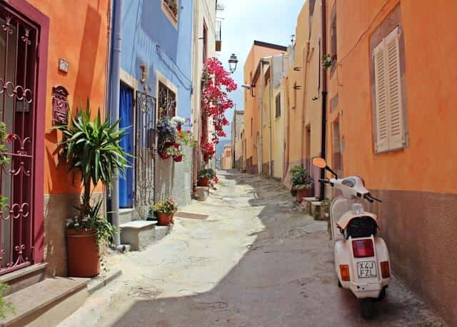 A white scooter in a small Italian street with colorful buildings on both sides.