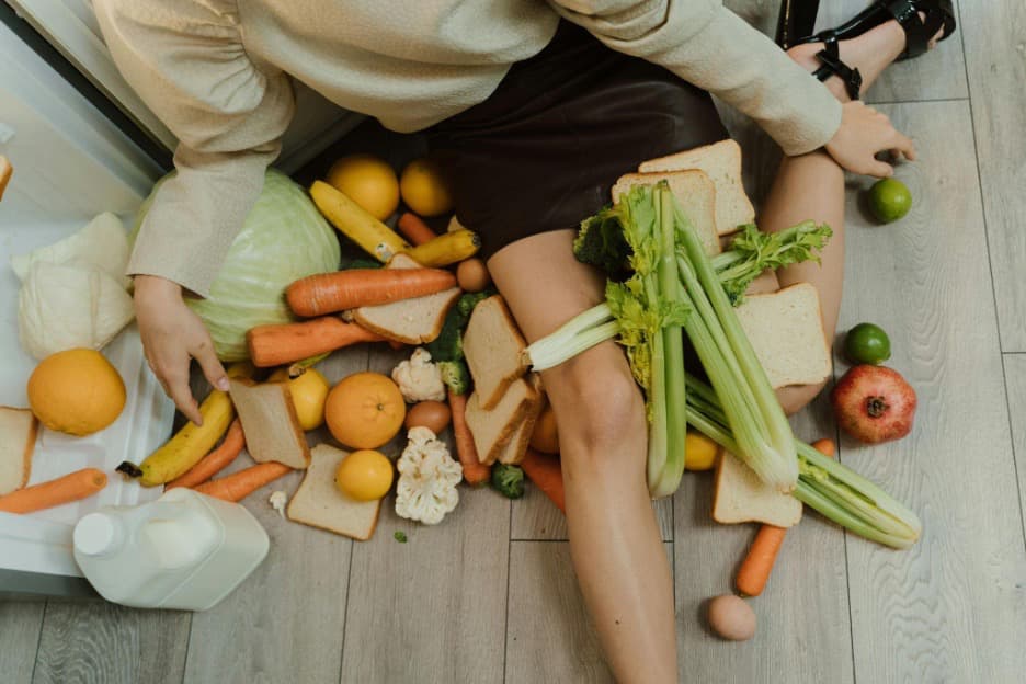 A woman sitting next to a fridge, with fruits and vegetables all around her.